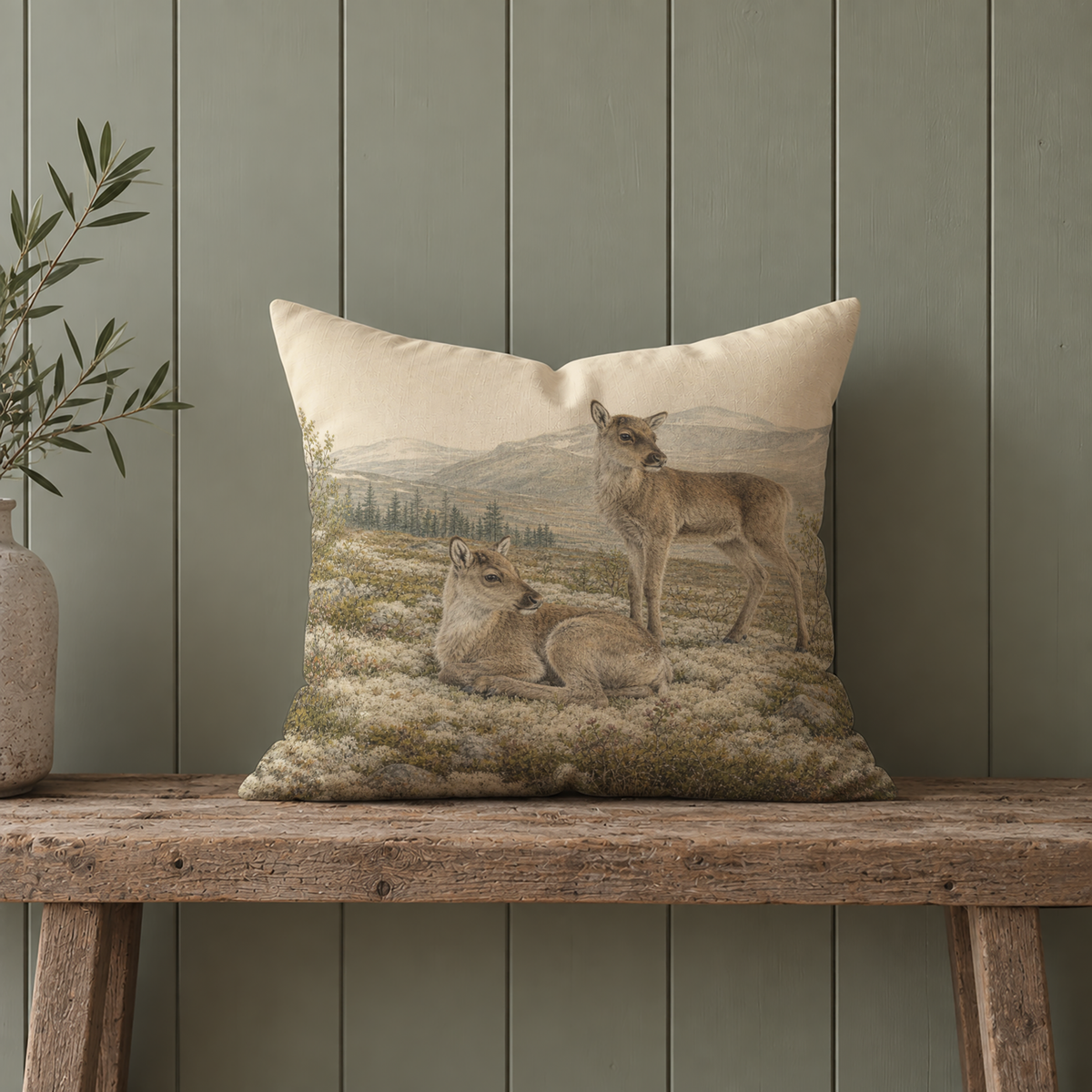 Reindeer decorative pillow cover with two young reindeer in a Nordic wilderness landscape, styled on a rustic wooden bench against a muted green paneled wall.